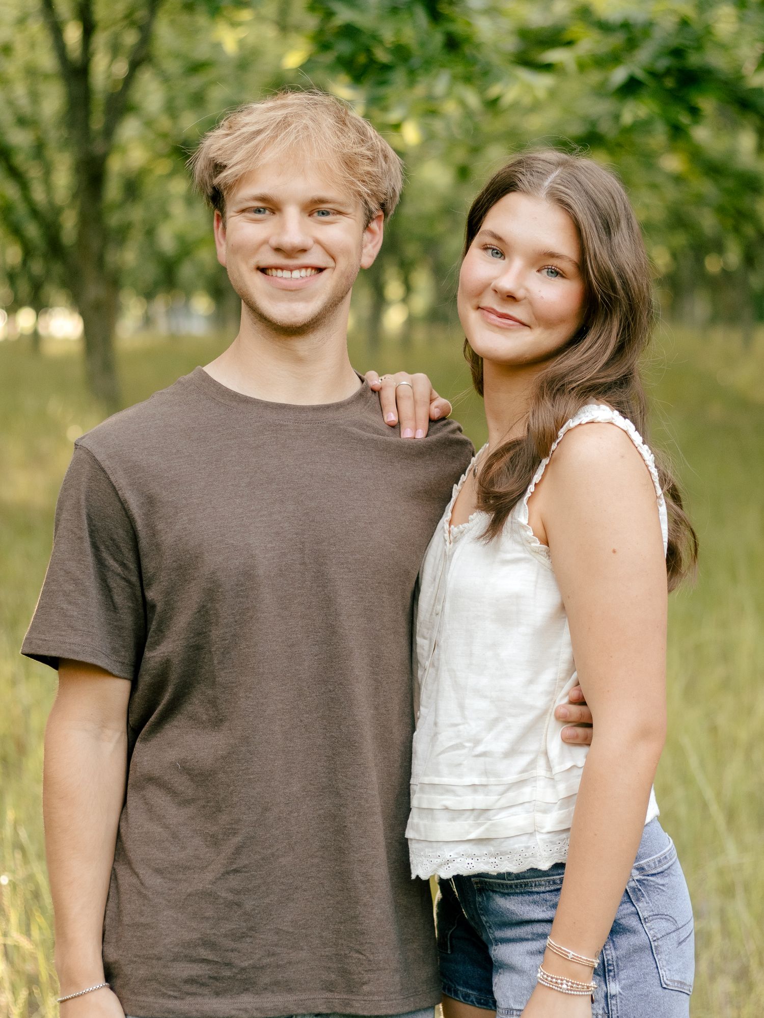 A smiling young man and woman stand close together with arms around each other in a sunlit grassy field with trees behind them.