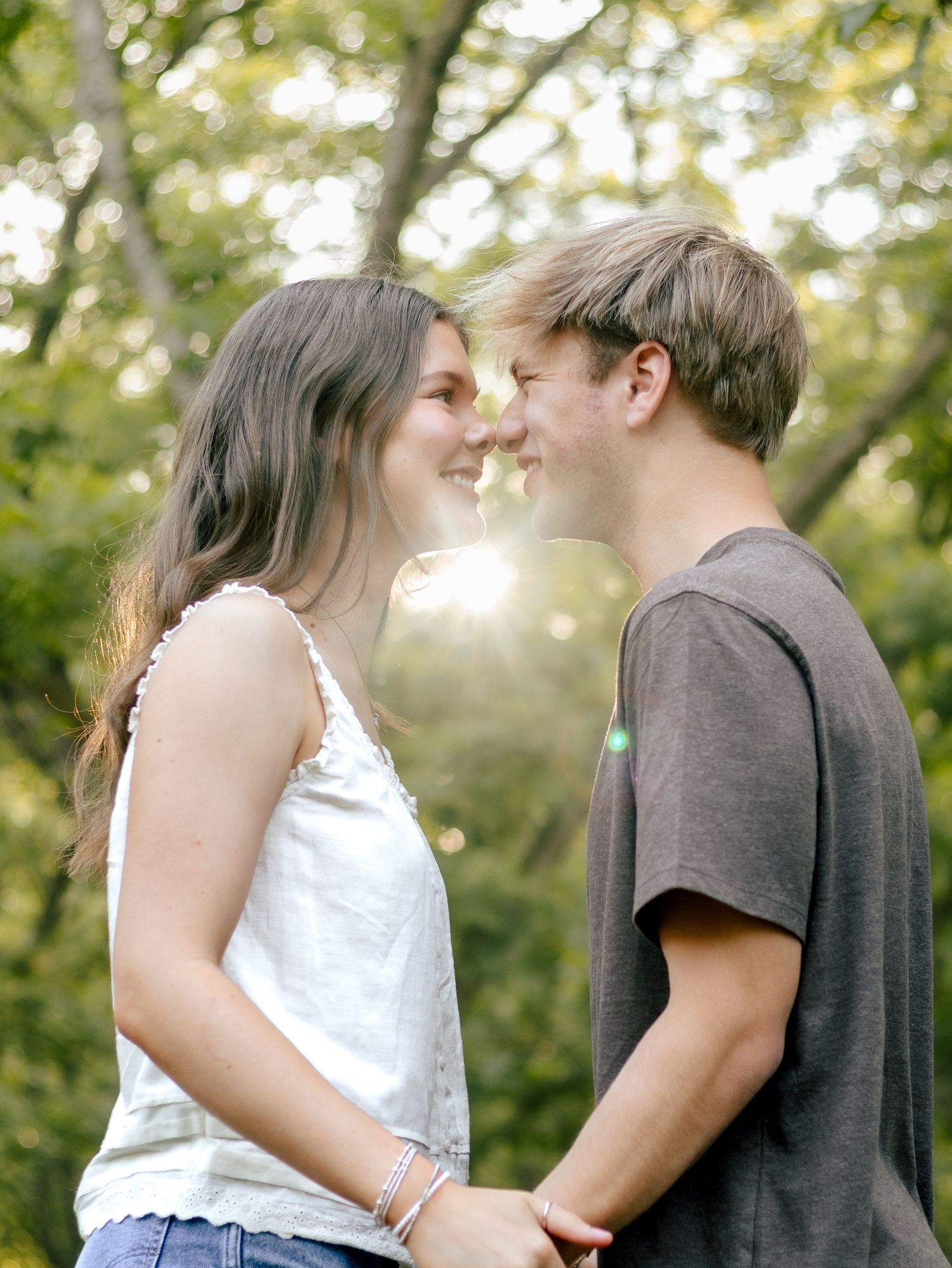 A smiling couple stands face to face with their noses touching in a sunlit park, surrounded by green trees and warm golden light.