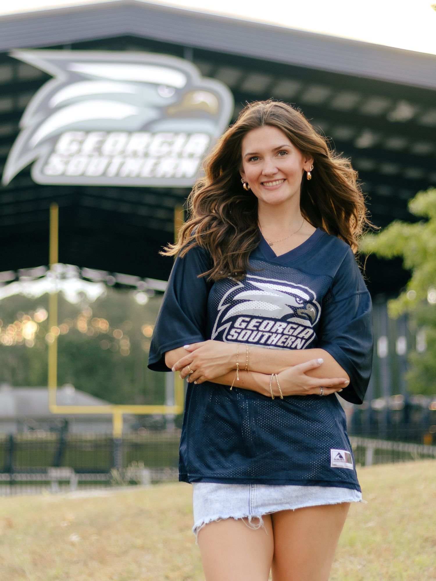 Woman with long wavy brown hair smiles in a Georgia Southern jersey and denim shorts in front of the Georgia Southern eagle stadium sign.