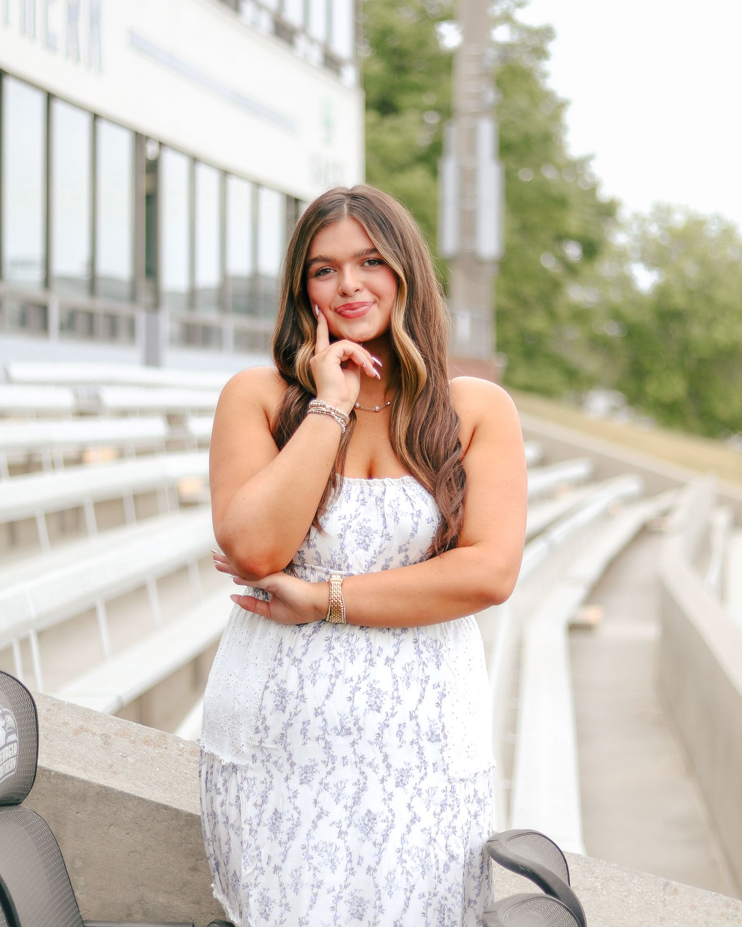 Woman with long wavy hair in a white floral sundress poses with her hand at her chin in front of empty stadium bleachers.