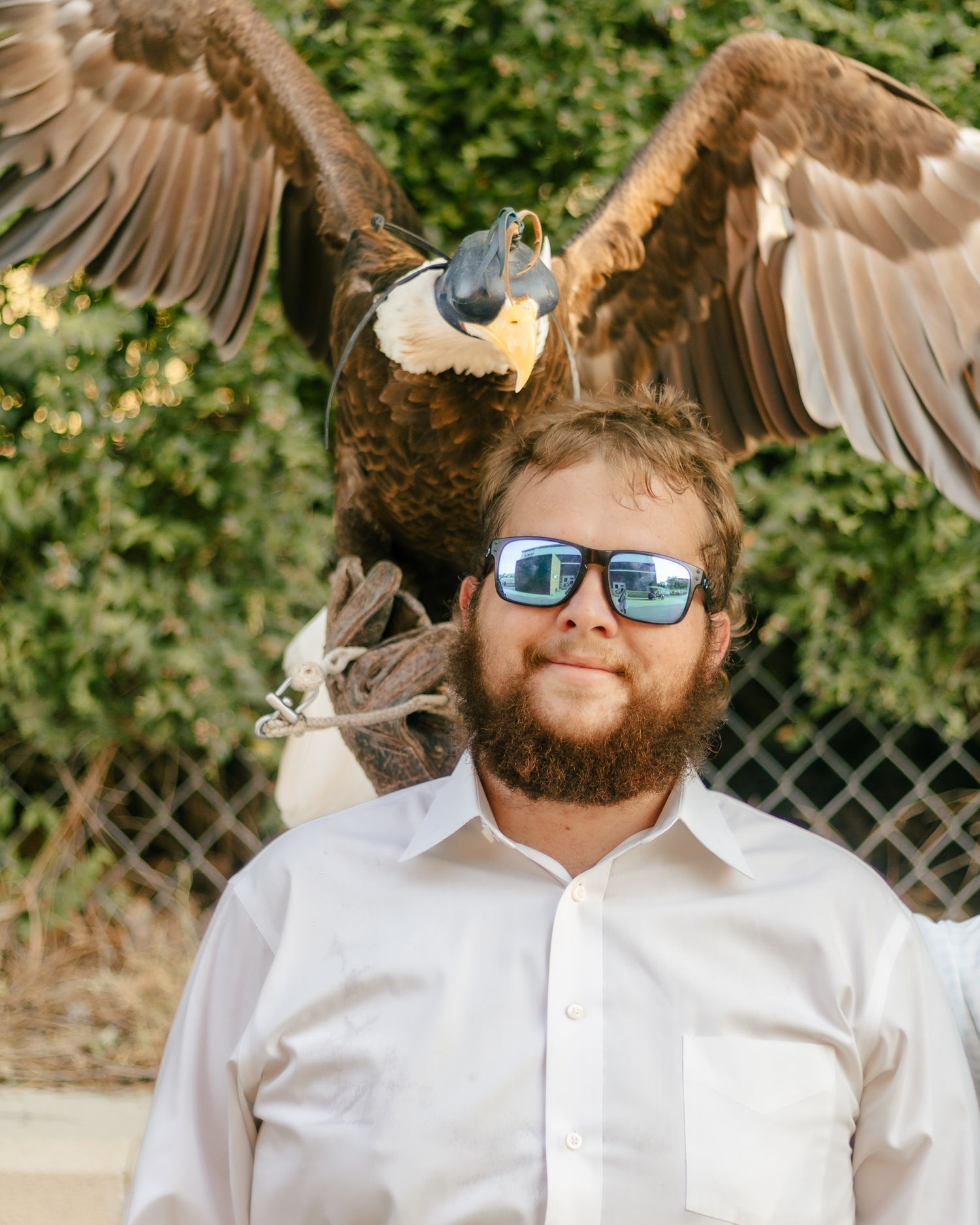 Bearded man in mirrored sunglasses and a white shirt stands outdoors as a large bald eagle perches on his shoulder with wings spread.