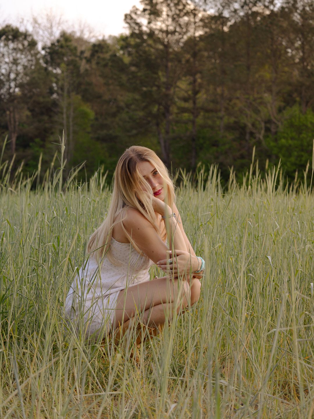 Woman with long blonde hair and red lipstick crouching in a tall grass field in a white dress with trees in the background.