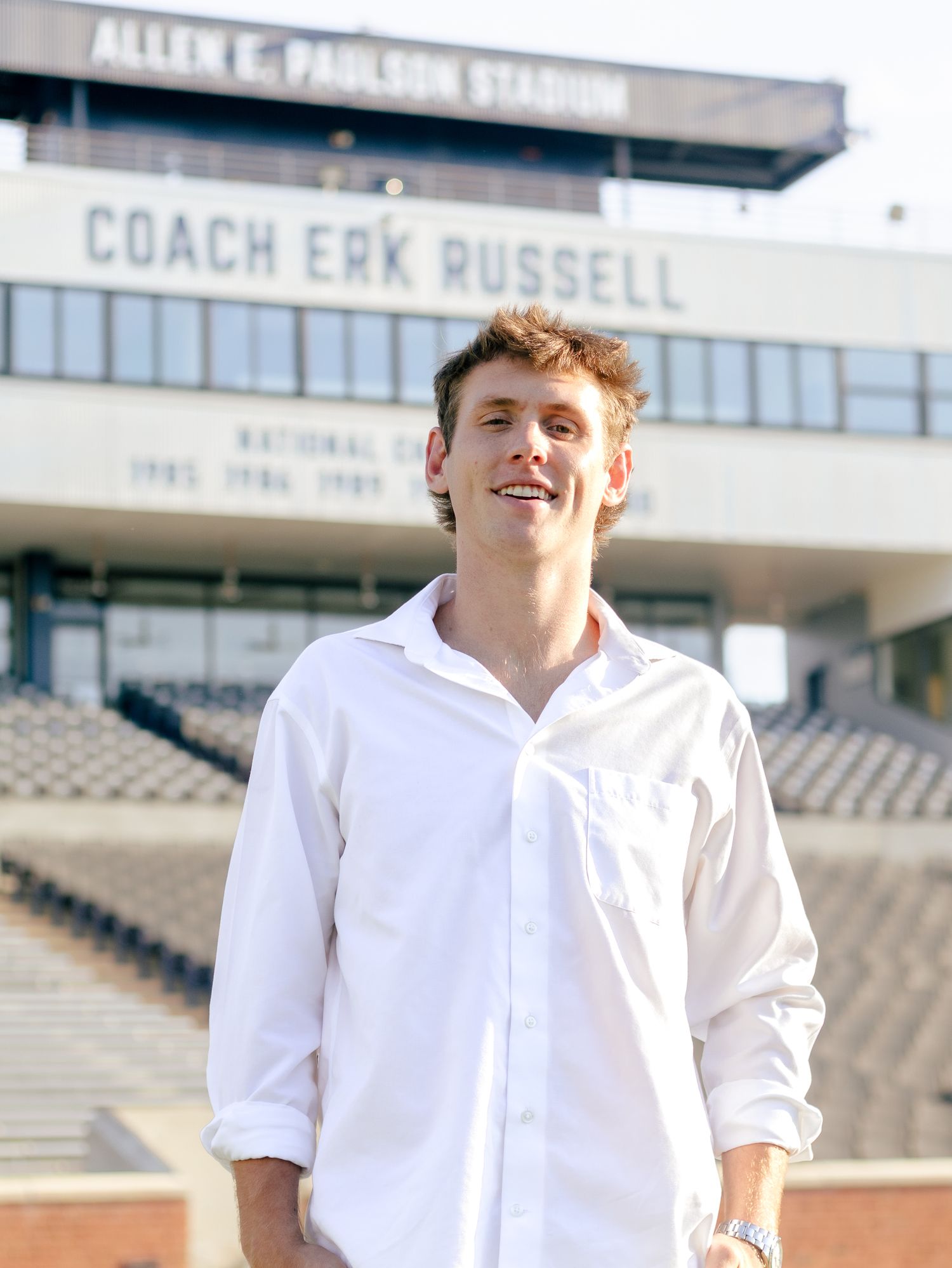 Smiling young man in a white button-up shirt stands in front of Allen E. Paulson Stadium's Coach Erk Russell press box and empty stands.