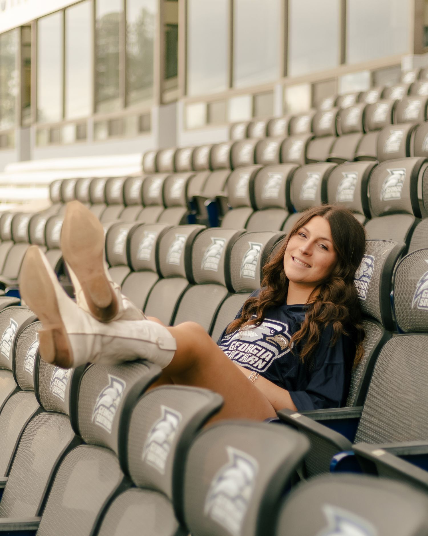Smiling woman in a Georgia Southern shirt lounges with her feet up in gray stadium seats featuring the Eagles logo.