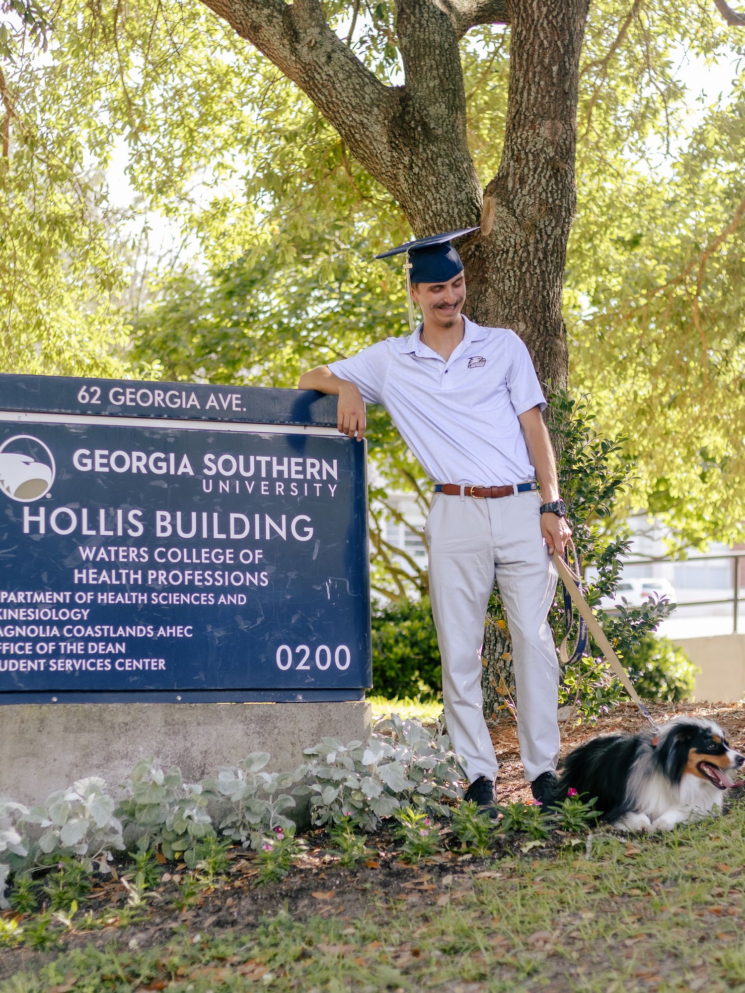Graduate in a mortarboard leans on the Hollis Building sign at Georgia Southern University while a black and white dog rests on the grass.