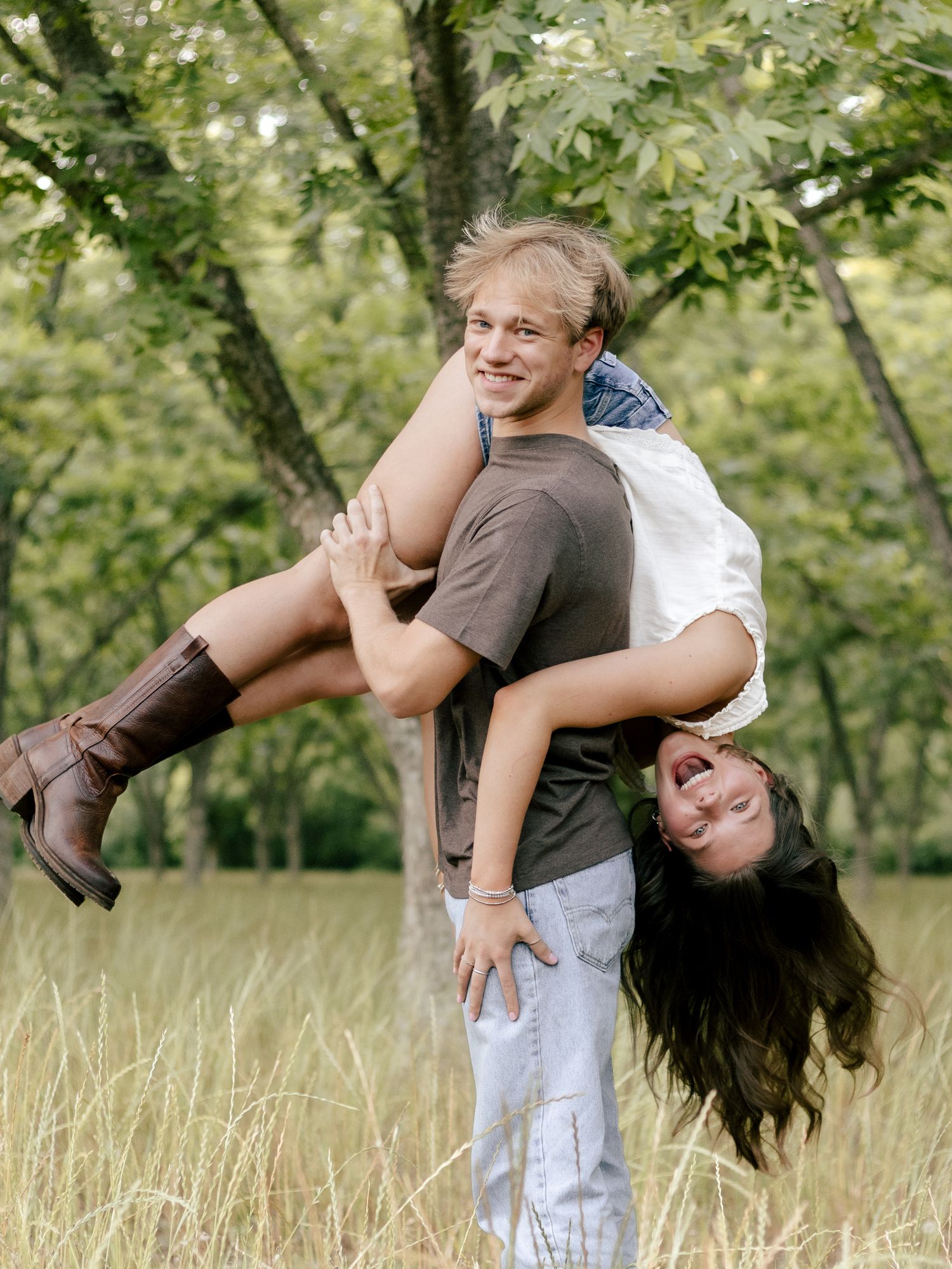 Smiling young man in a brown t-shirt playfully lifts a laughing woman with long dark hair over his shoulder in a sunny grassy field.
