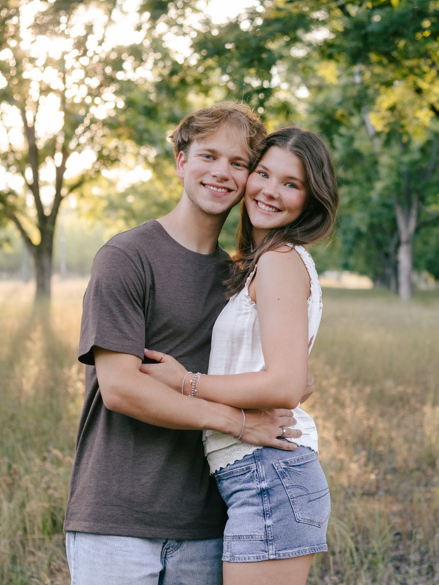 Smiling young couple embracing outdoors in a sunlit grassy field surrounded by trees during a golden hour portrait session.