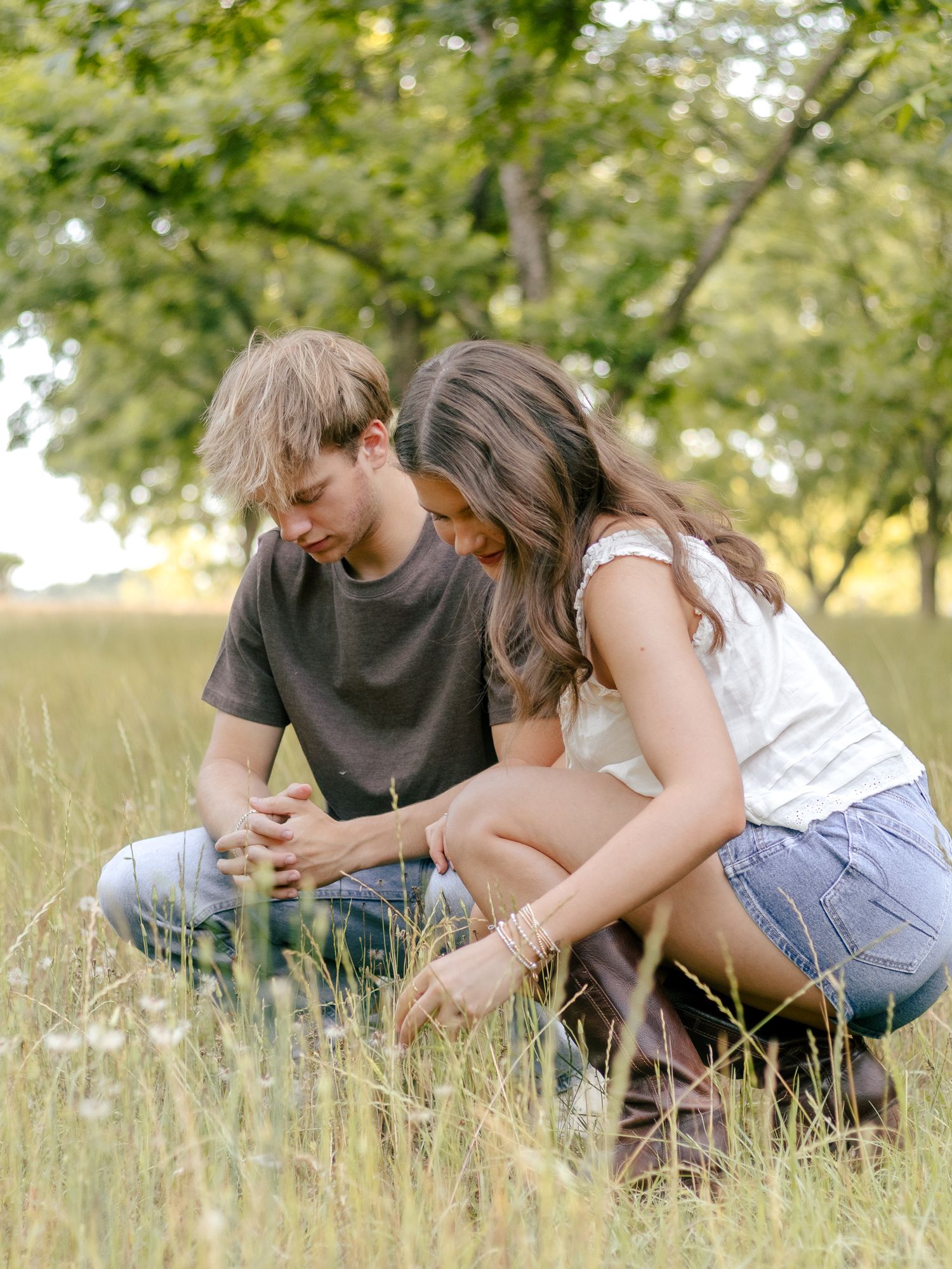 Young man and woman sitting close together in a tall grassy field, heads bowed and hands clasped, under green trees.