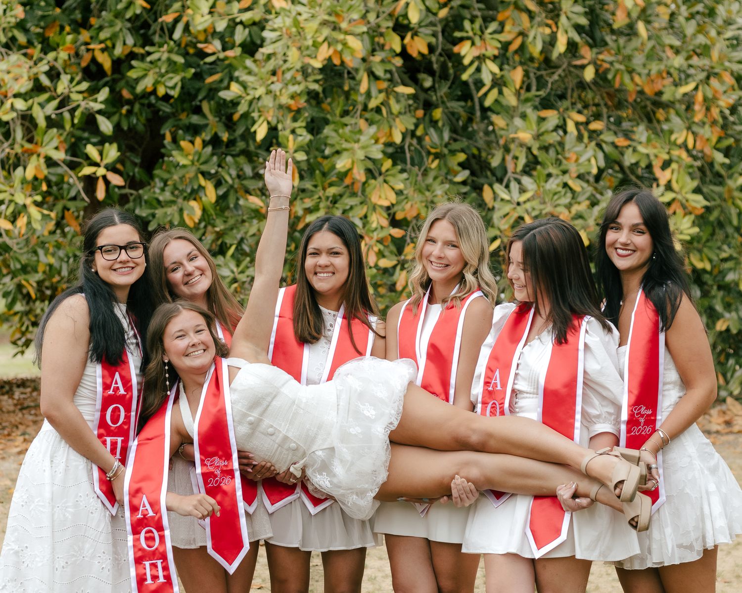 Group of smiling young women in white dresses and red sashes lift a woman across their arms outdoors in front of a leafy green tree.