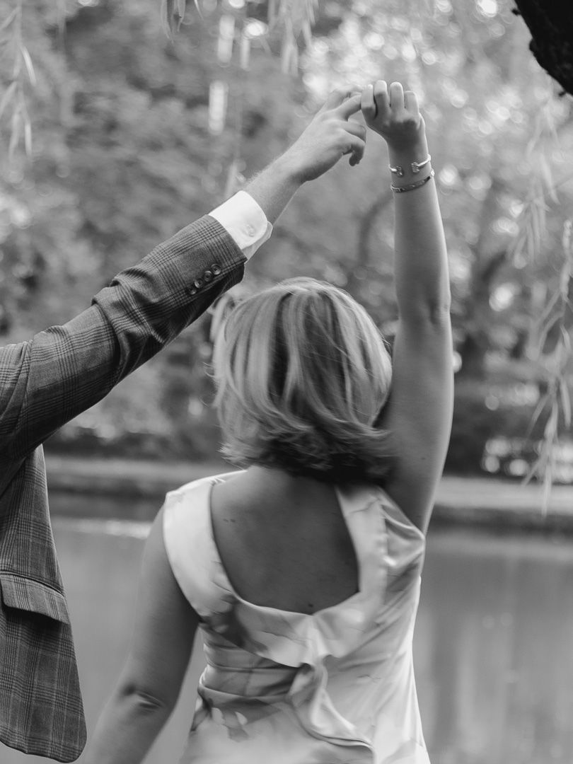 Black and white photo of a woman in an elegant dress twirling with her raised hand held by a partner beside a tranquil lake.