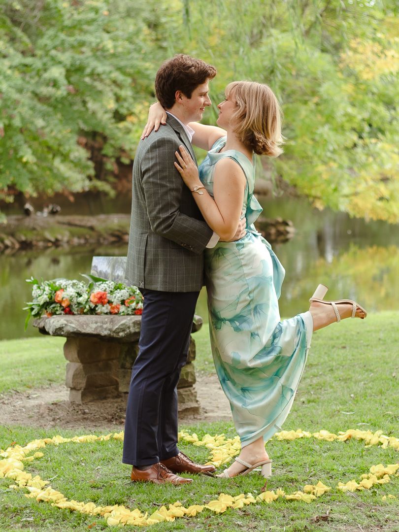 Couple embracing in a park beside a pond, the woman in a green dress kicking up her heel, with flower petals and a bouquet nearby.