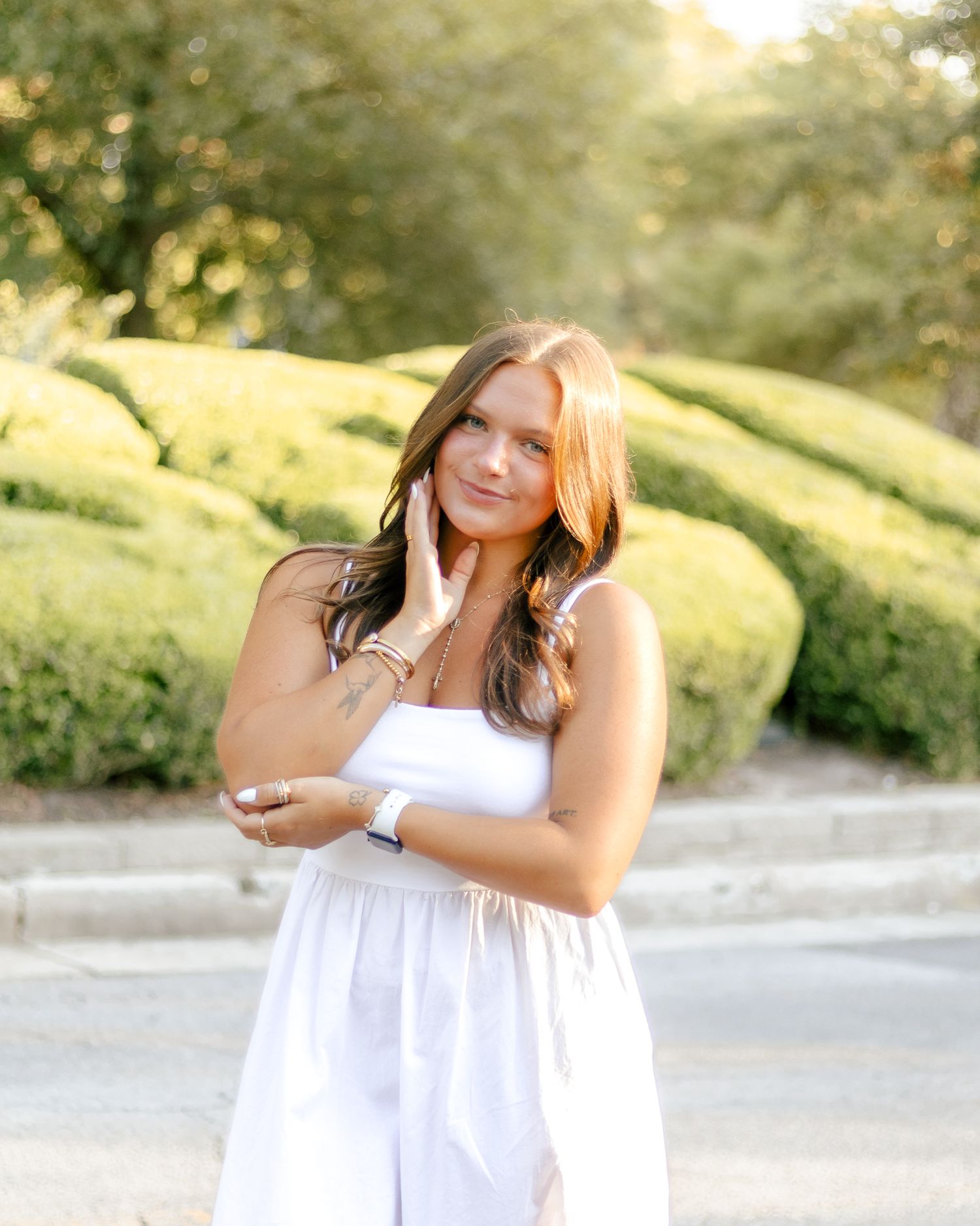 Smiling woman with long auburn hair in a white sleeveless dress poses outdoors with a hand near her face beside neatly trimmed green hedges.