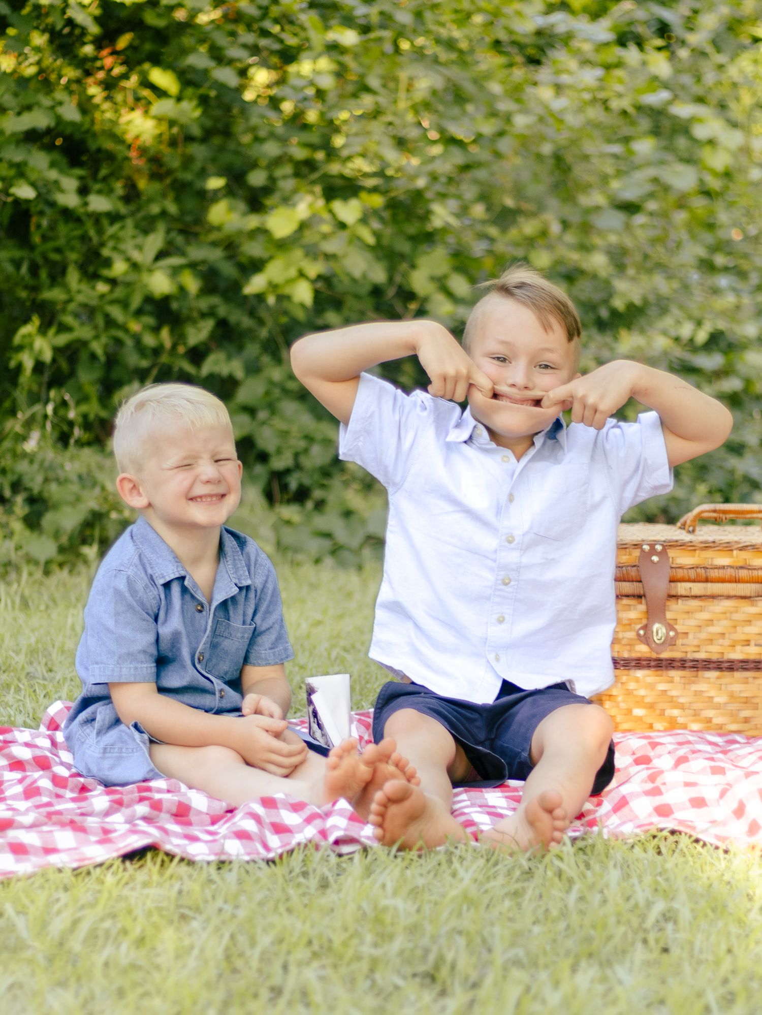 Two smiling young boys sit barefoot on a red checkered picnic blanket beside a wicker basket, making silly faces on a sunny grassy lawn.