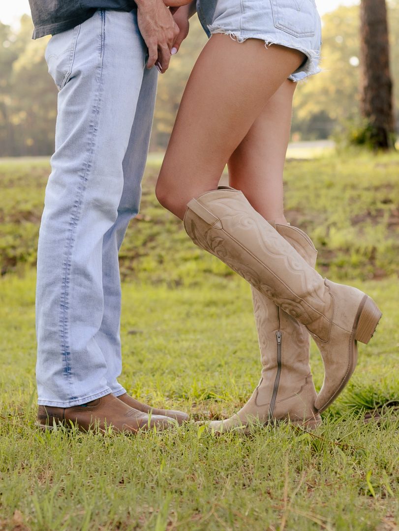 Couple standing close on green grass, her in tan cowboy boots and denim shorts, him in light-wash jeans and brown boots.