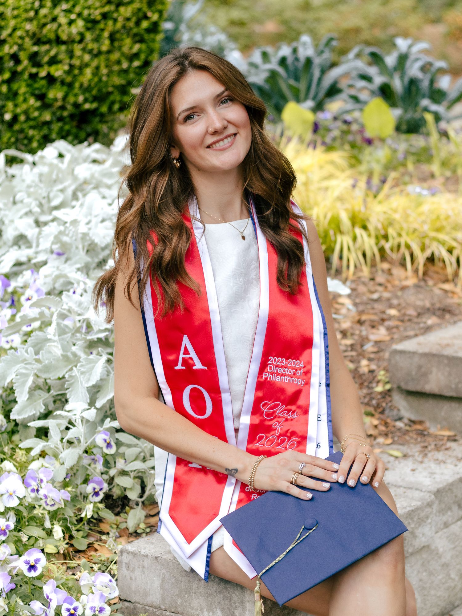 Smiling graduate in a red and white Alpha Omicron stole sits on stone steps holding a navy graduation cap beside a lush flower garden.