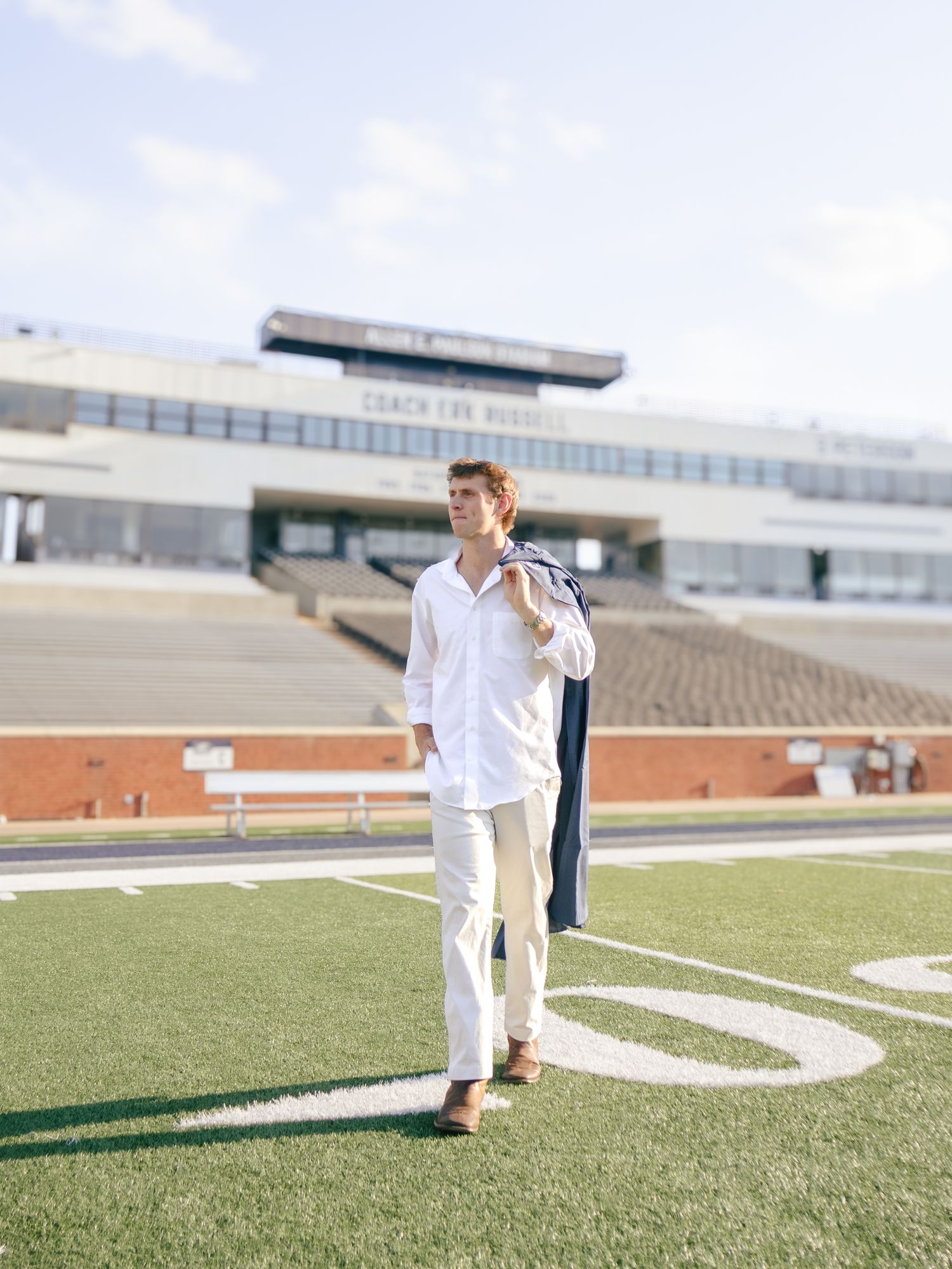 Young man in white linen shirt and pants holds a navy jacket over his shoulder on a football field with stadium bleachers behind him.