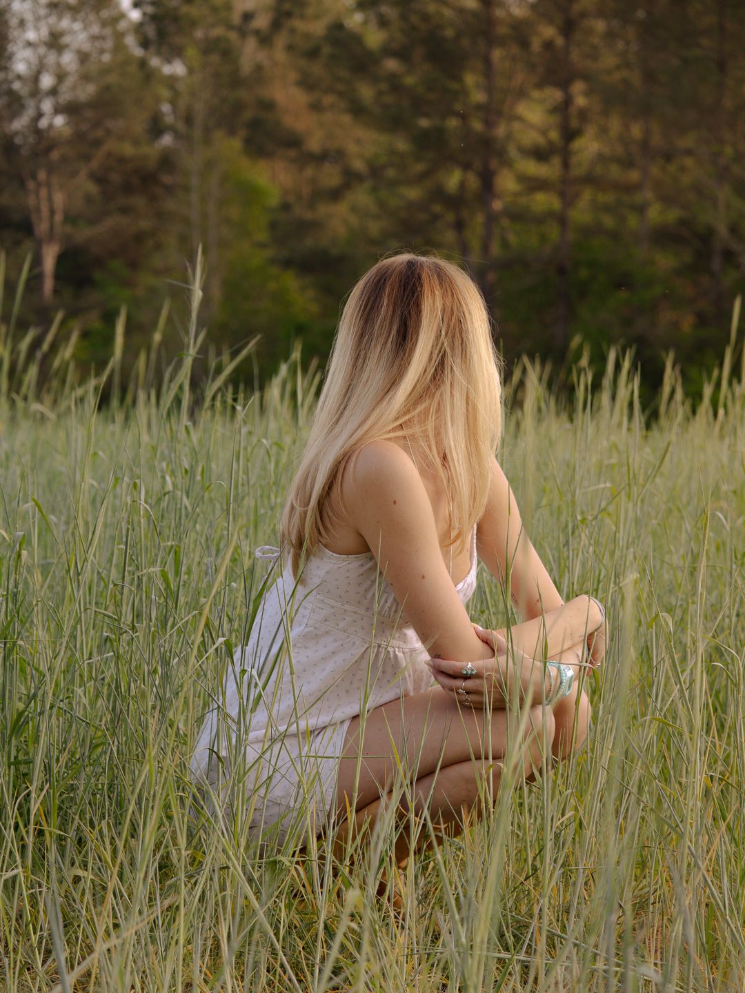 Woman with long blonde hair in a white dress crouching in tall green grass, facing a forest at golden hour.