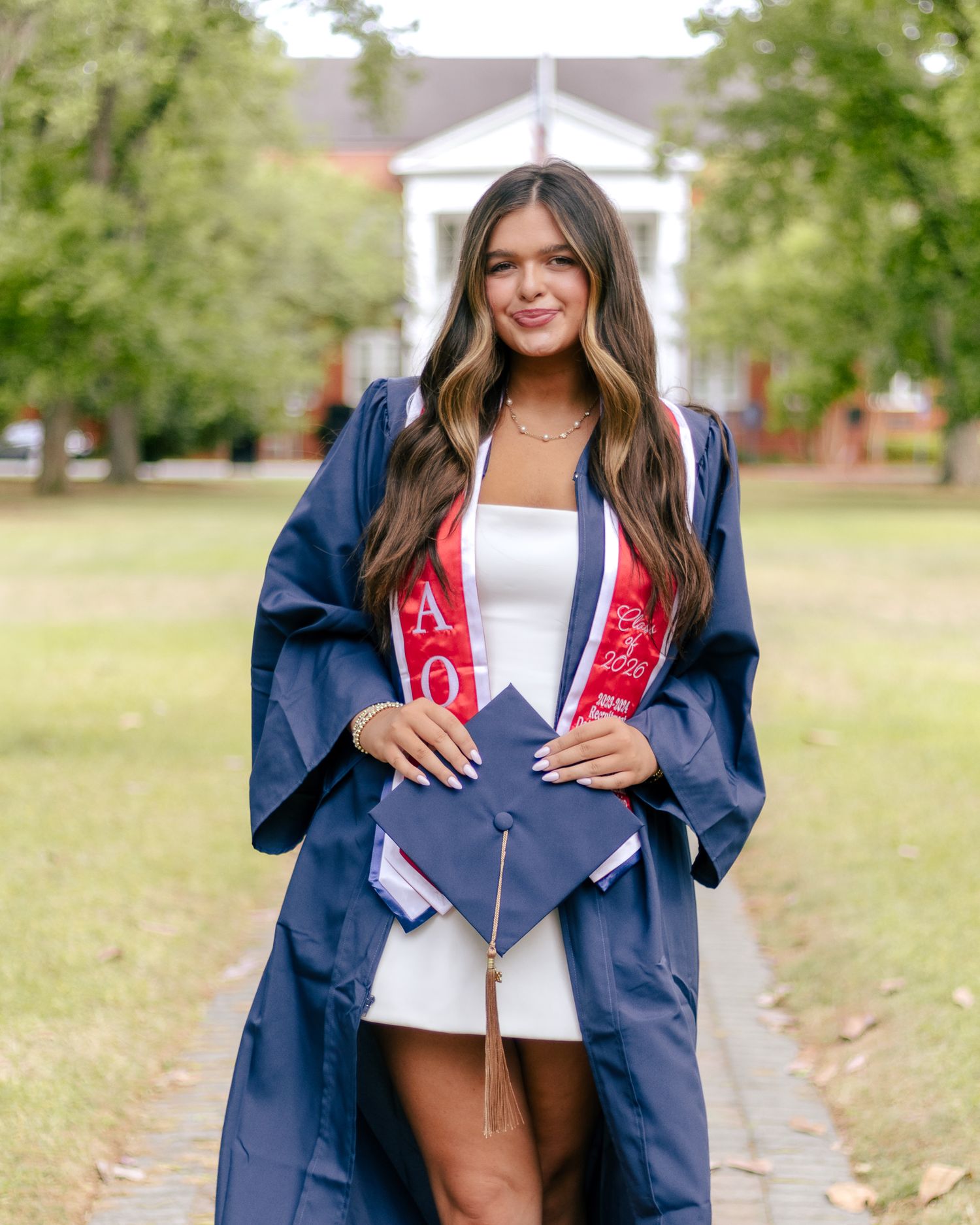 Smiling graduate in a navy cap and gown with red sorority stole holds her mortarboard on a tree-lined campus lawn.