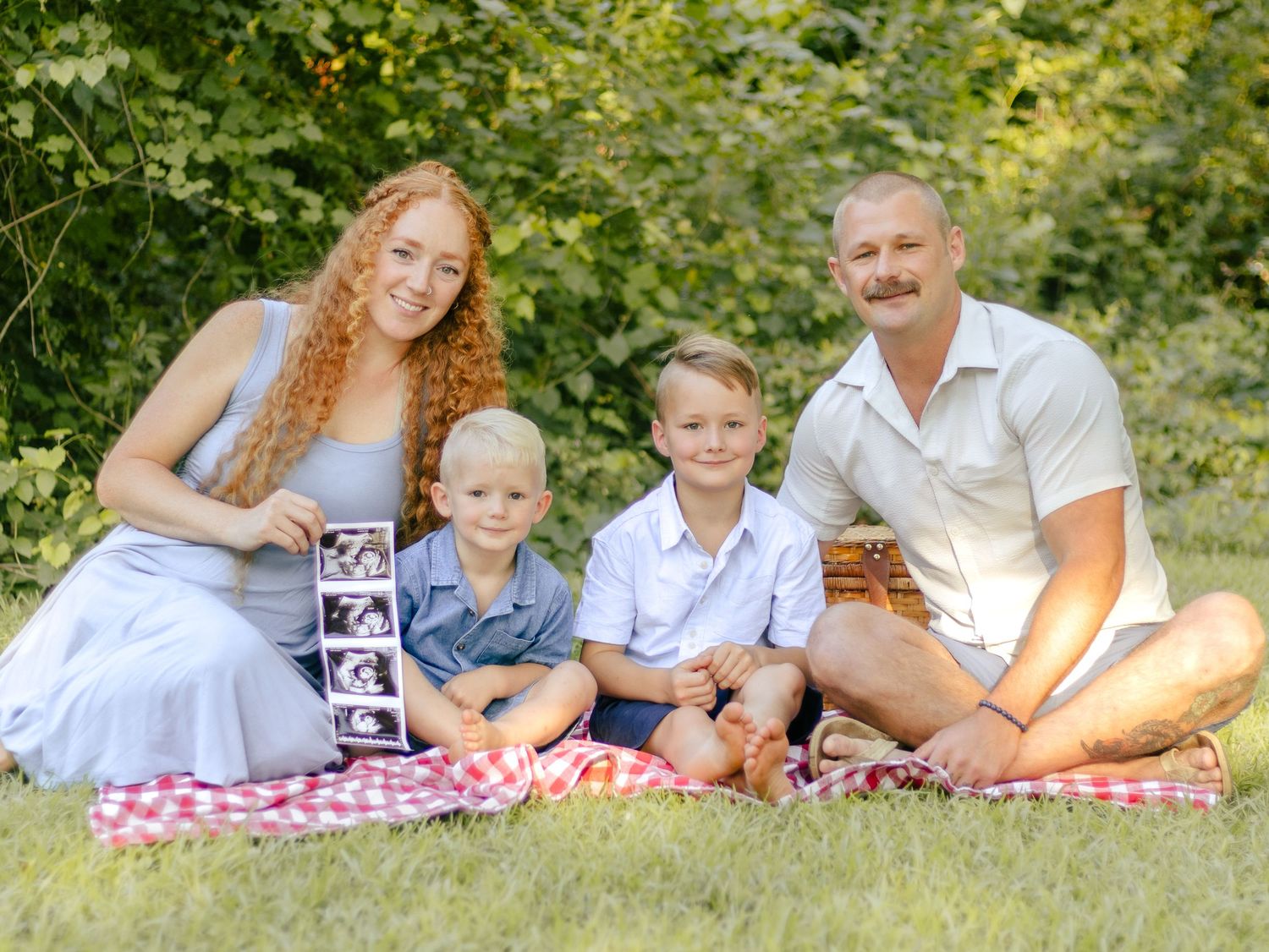 Family of four with two young boys sitting on a red checkered picnic blanket on grass, holding ultrasound photos to announce a new baby.