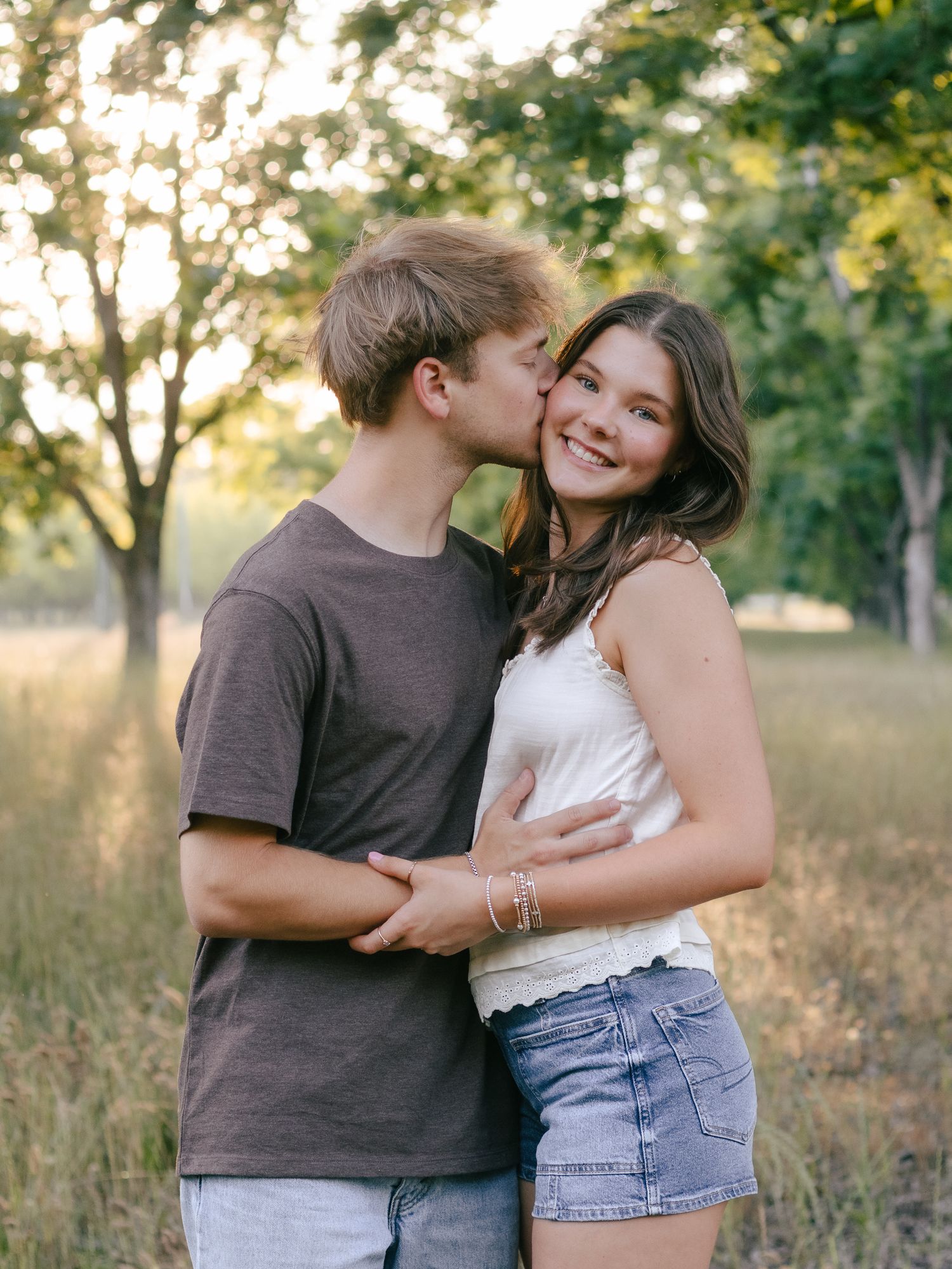 A young man in a brown t-shirt kisses the cheek of a smiling young woman in a white top and denim shorts in a sunlit grassy field.