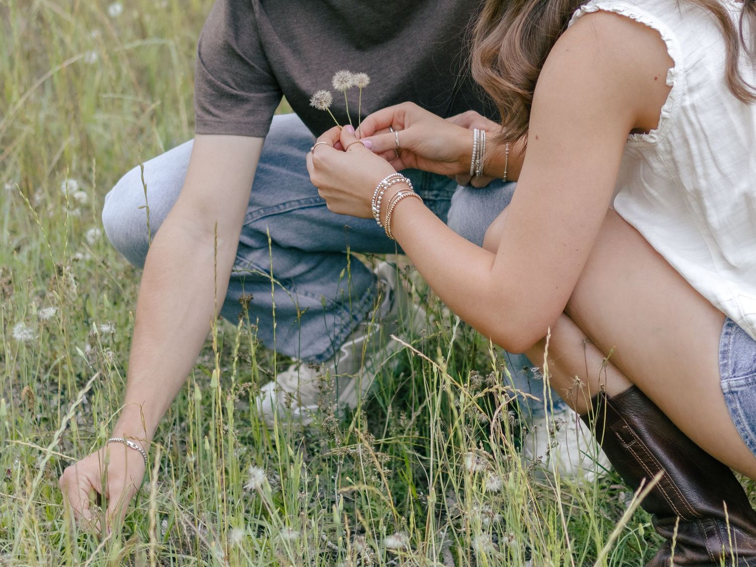 Two people crouch in a sunlit meadow of tall grass and white wildflowers, gently picking small blooms together.