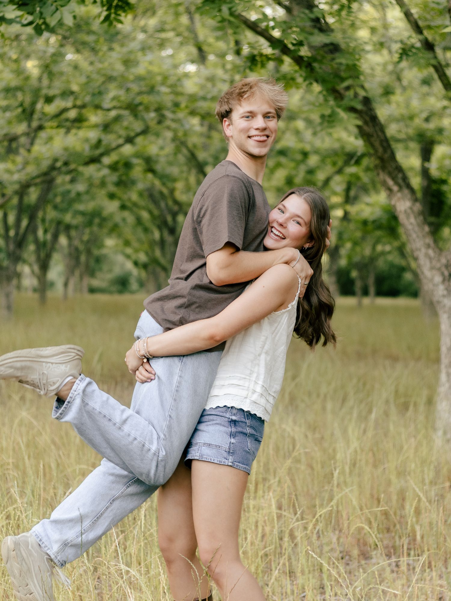 A smiling young man in a gray t-shirt lifts a laughing young woman in a white top and denim shorts in a grassy field with trees.