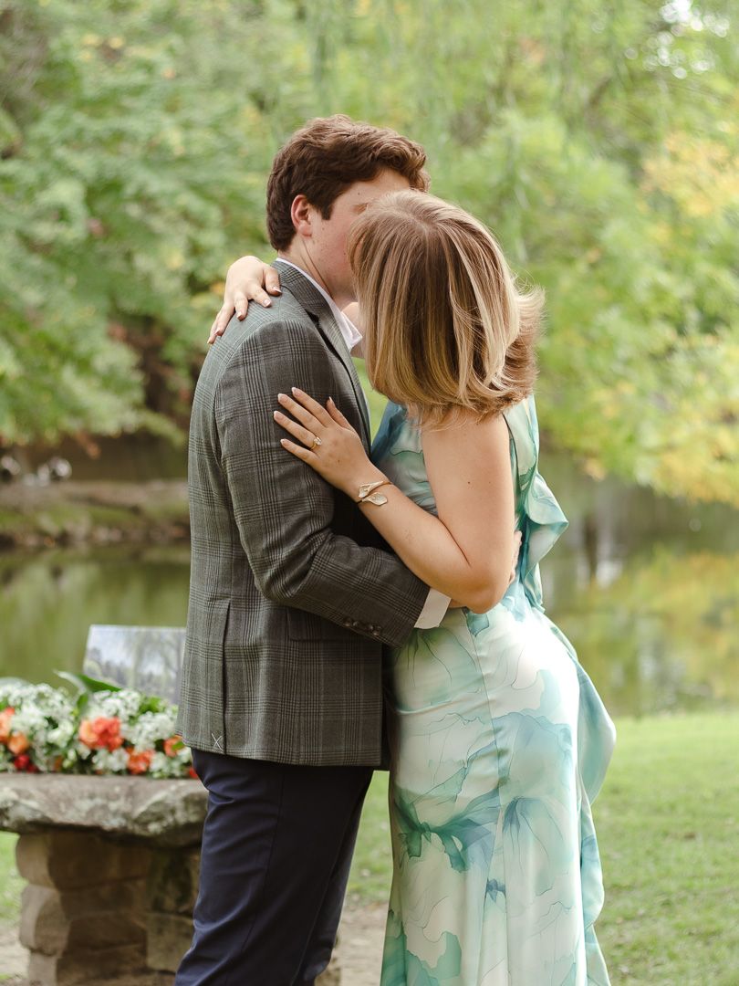 A newly engaged couple hugs tightly beside a stone pedestal with flowers near a calm lake.