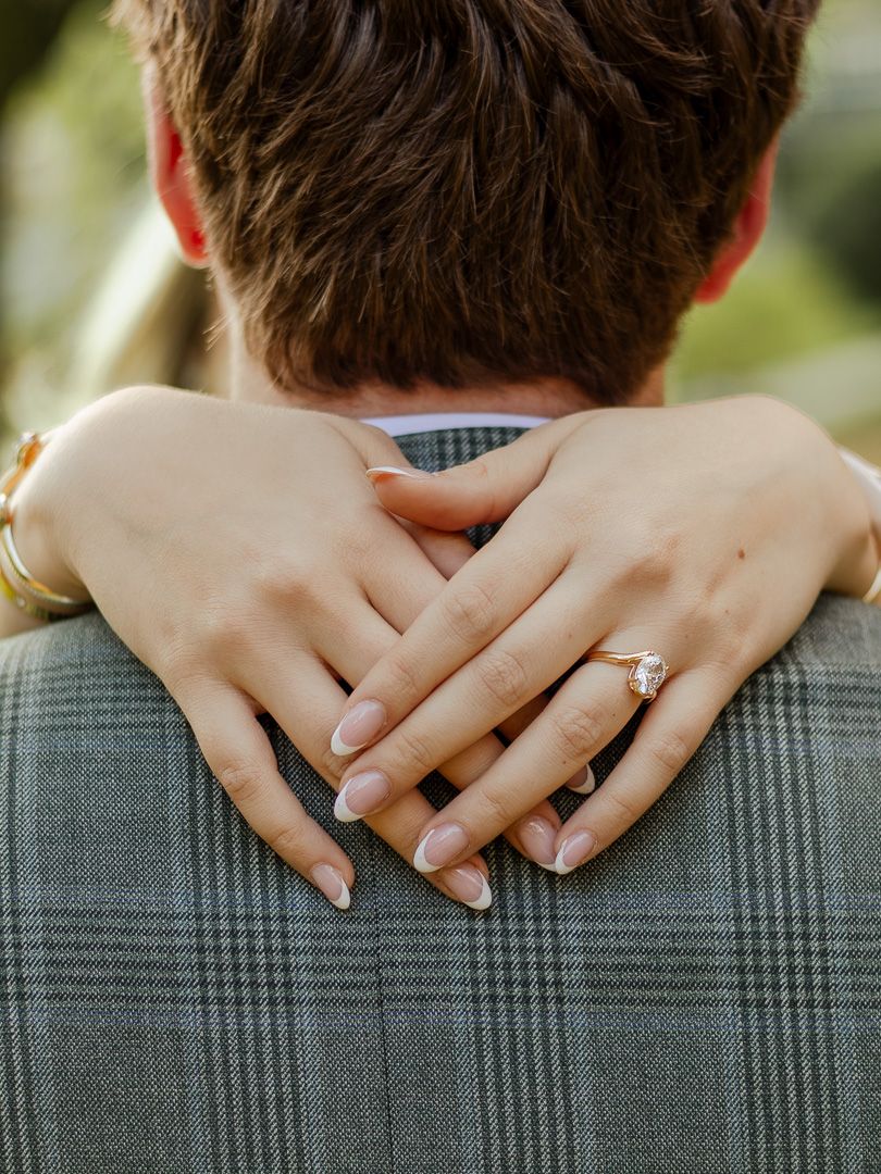 Woman's hands with a diamond engagement ring wrapped around the neck of a man in a gray plaid suit jacket outdoors.