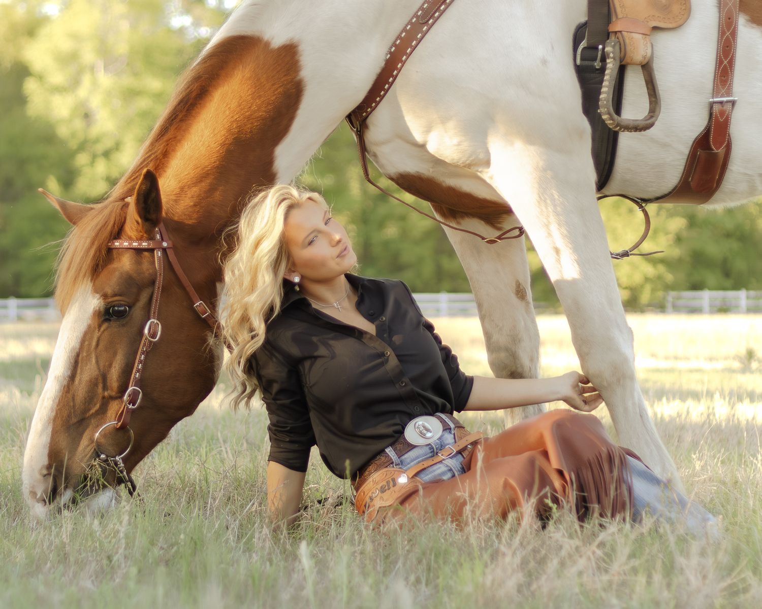 Blonde woman in a dark shirt and boots reclines in a sunny grassy field beside a grazing brown and white paint horse in a western saddle.
