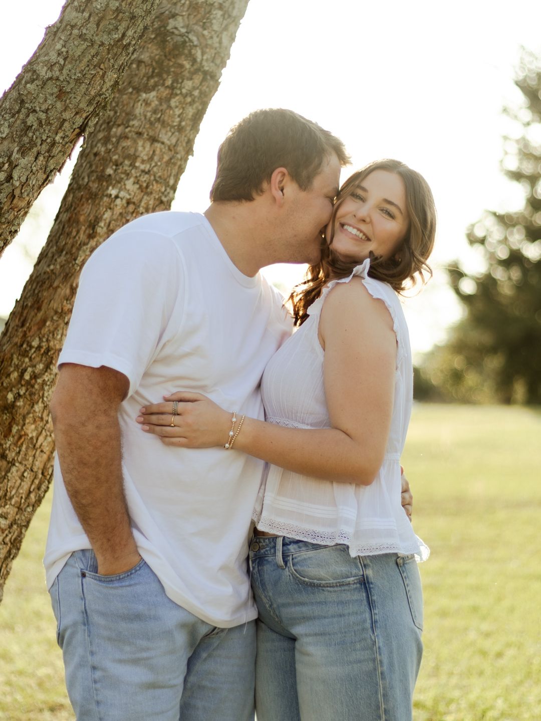 Smiling couple in jeans and white tops embrace as he kisses her cheek beside a tree in a sunny, grassy park.