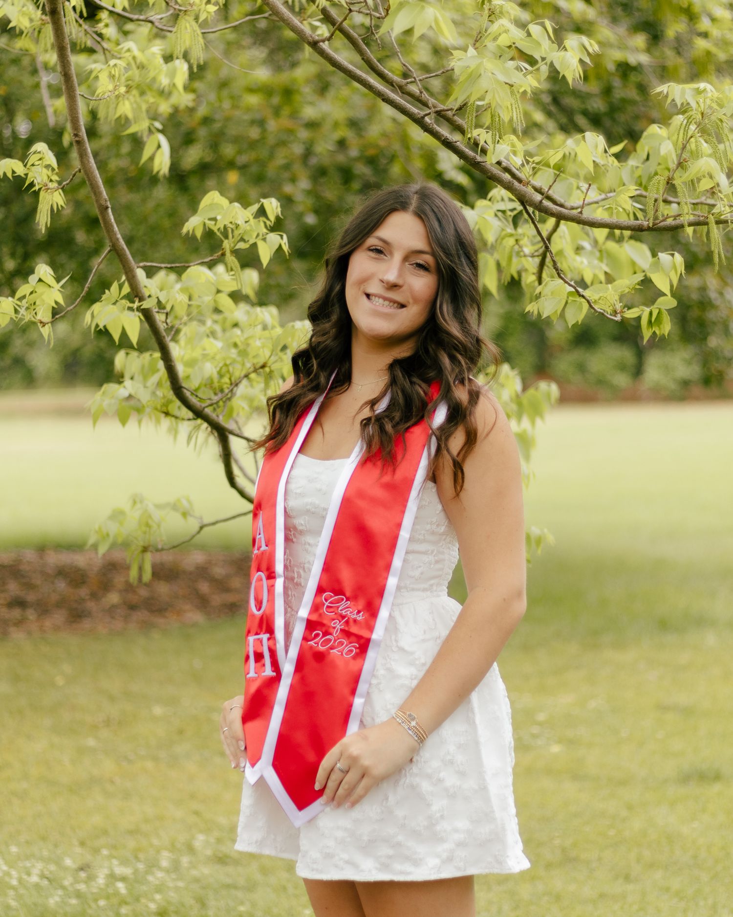 Smiling graduate in a white dress and red graduation stole poses outdoors on a green lawn beneath leafy tree branches.