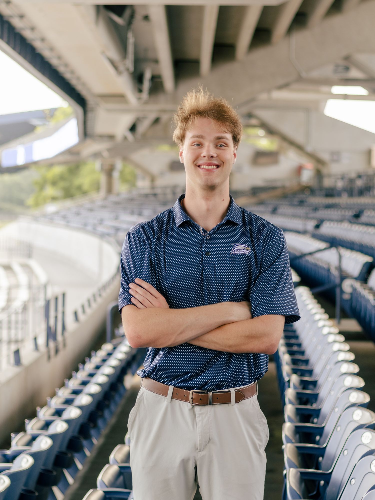 Smiling young man in a navy polo and khaki pants stands with arms crossed among rows of empty blue stadium seats under a concrete overhang.