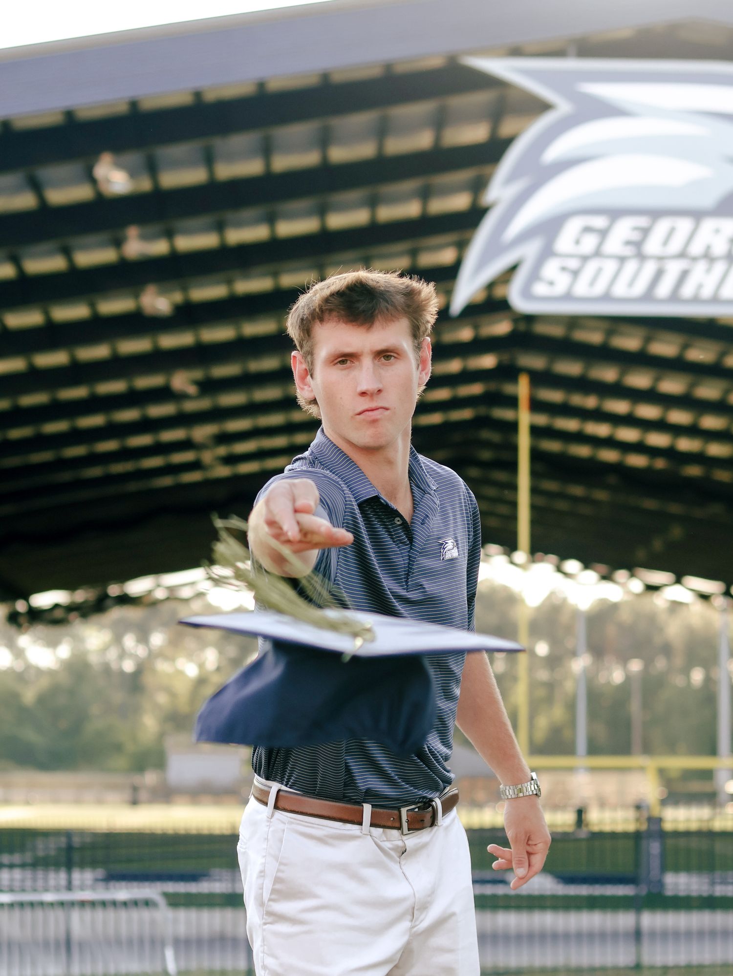 Young man in a polo shirt and khaki shorts tosses a paper airplane in front of a covered stadium stand with Georgia Southern signage.