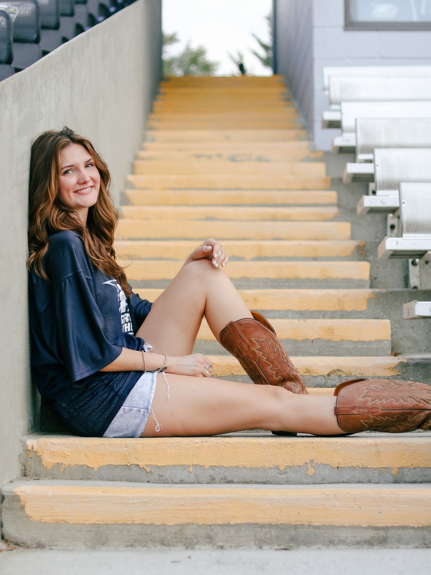 Smiling young woman with long wavy hair sits on concrete stadium steps wearing a navy graphic tee, denim shorts and brown cowboy boots.