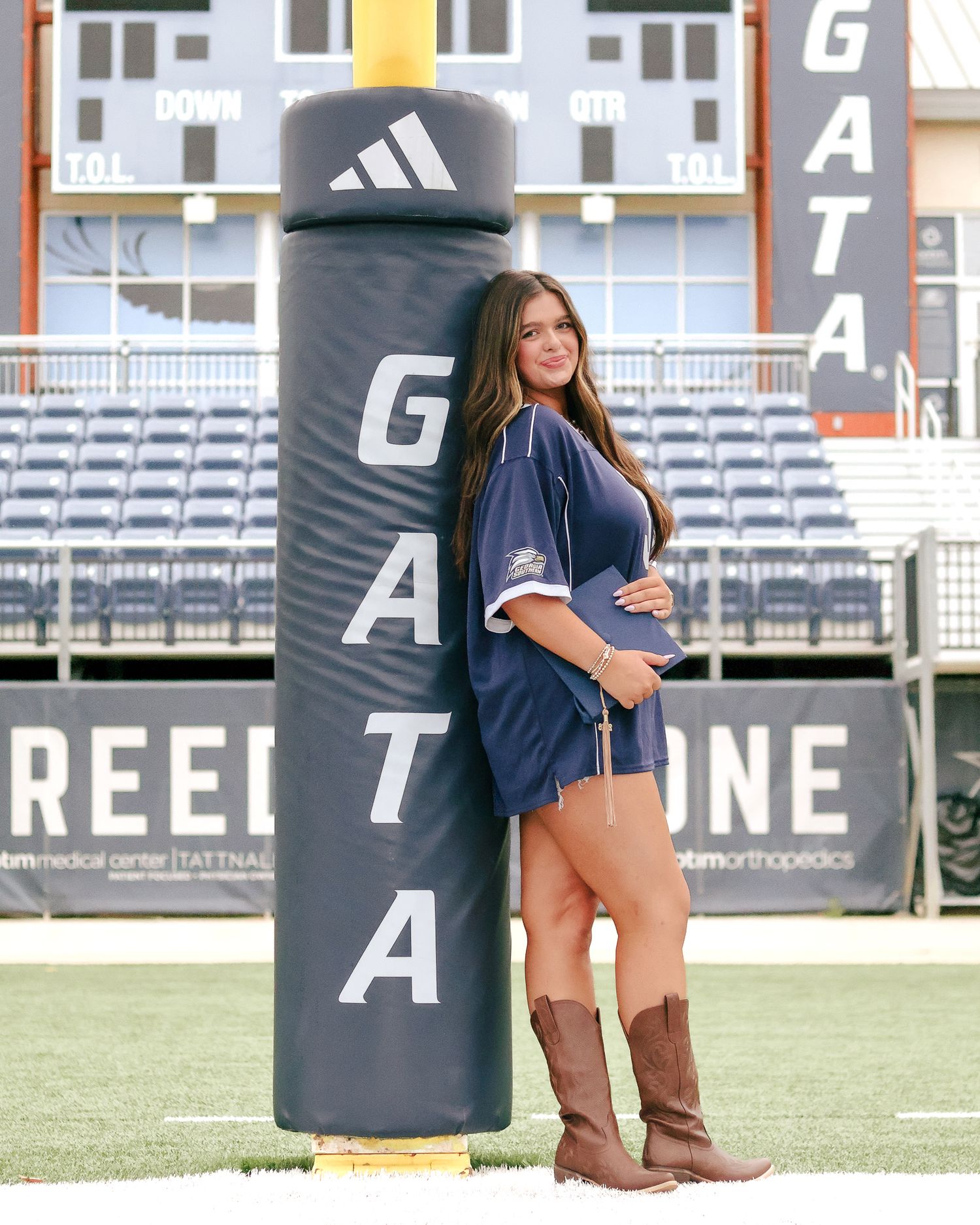 Young woman in a navy jersey dress and tan cowboy boots poses against a padded goalpost at a football stadium with bleachers behind her.