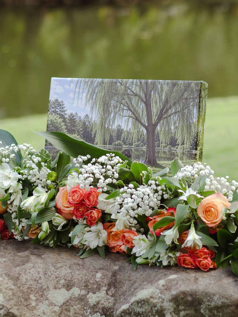 Peach roses, white alstroemeria and baby's breath arranged on a stone ledge beside a framed photo of a willow tree by a pond.