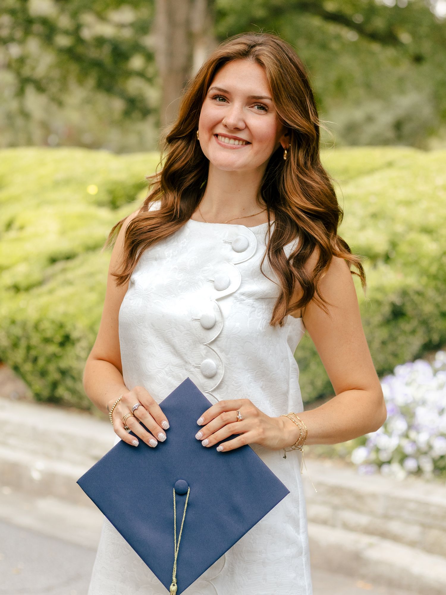 Smiling young woman with long wavy auburn hair in a white button-detail dress holding a navy graduation cap outdoors near green shrubs.