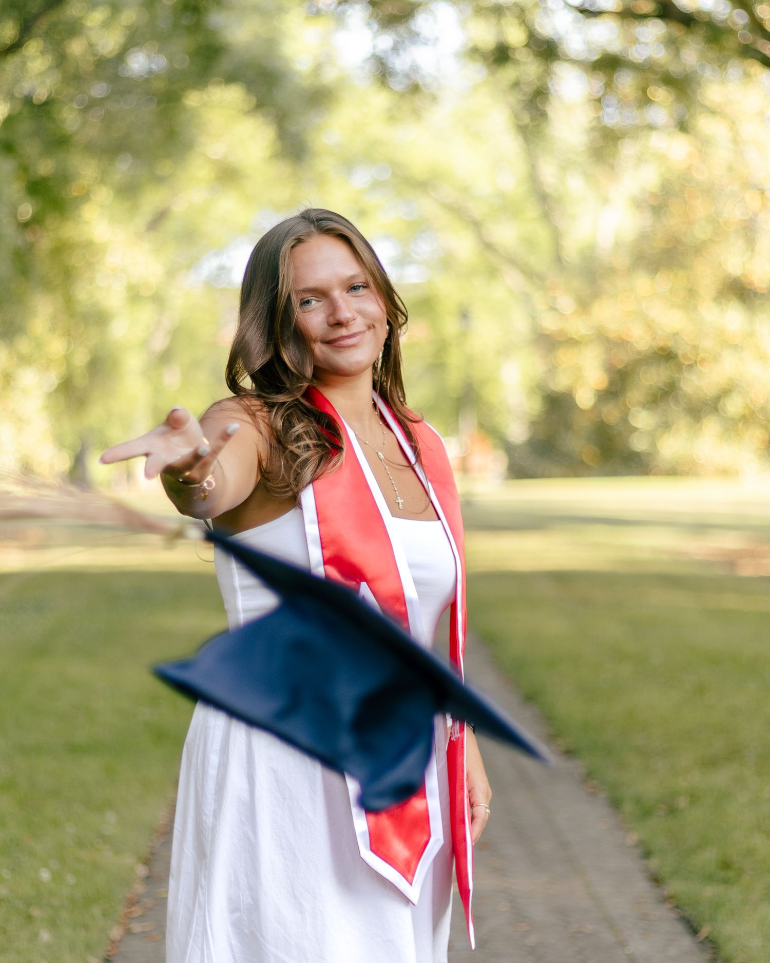 Smiling graduate in a white dress and red stole tosses her navy blue graduation cap in the air on a tree-lined path.