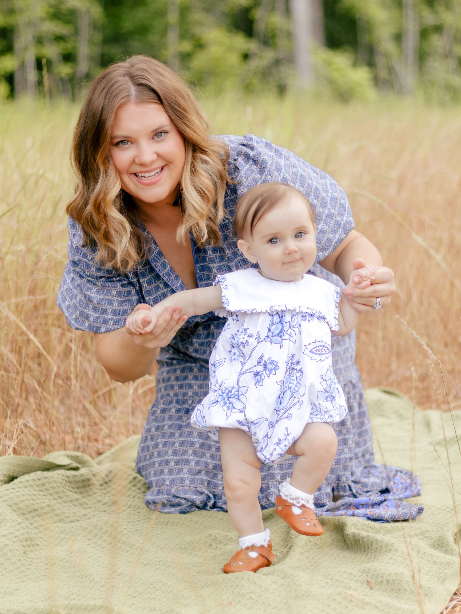 Smiling woman in a blue patterned dress kneels on a green blanket in a golden field, holding the hands of a toddler in a floral romper.