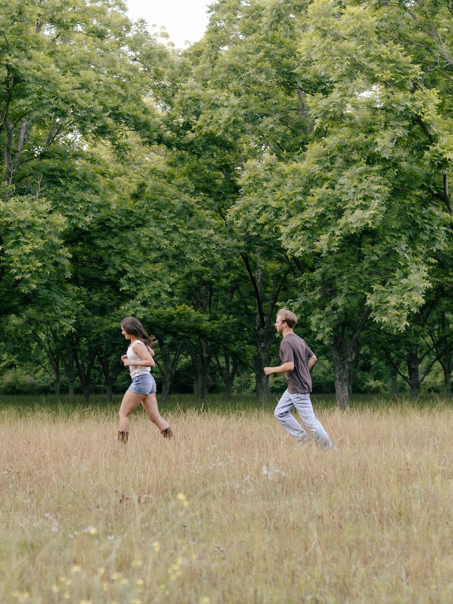Two people jog side by side through a sunlit field of tall dry grass, with lush green trees in the background.