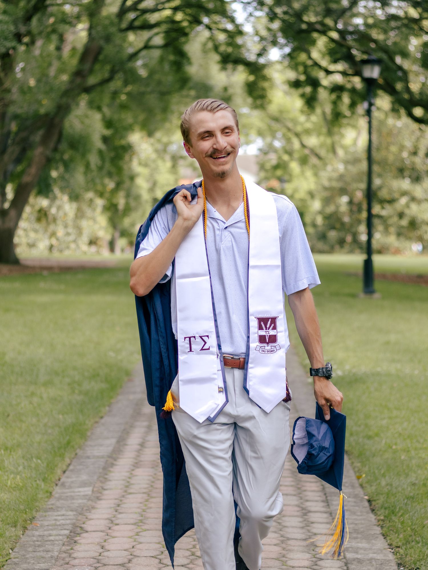 Smiling graduate in a white Greek letter stole carries his navy gown and cap while walking along a brick path on a sunny campus.
