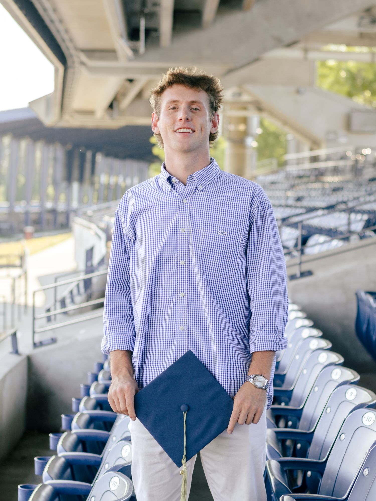 Smiling graduate in a blue checkered button-up shirt holding a navy mortarboard cap while standing among stadium seats under a grandstand.