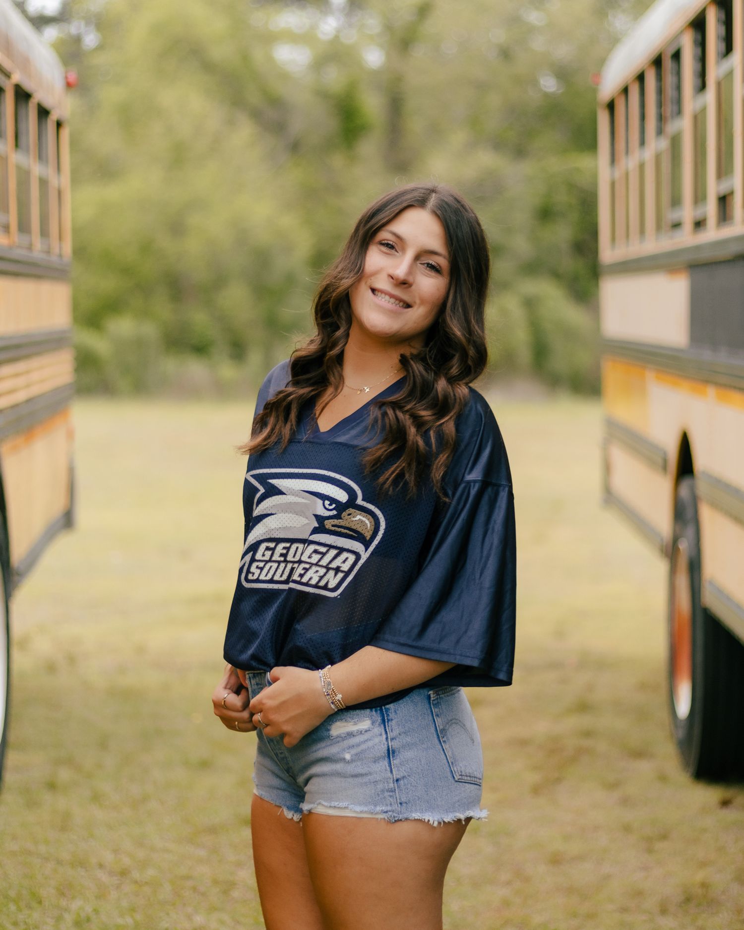 Smiling young woman with long wavy brown hair in a Georgia Southern jersey and denim shorts posing between two yellow school buses.