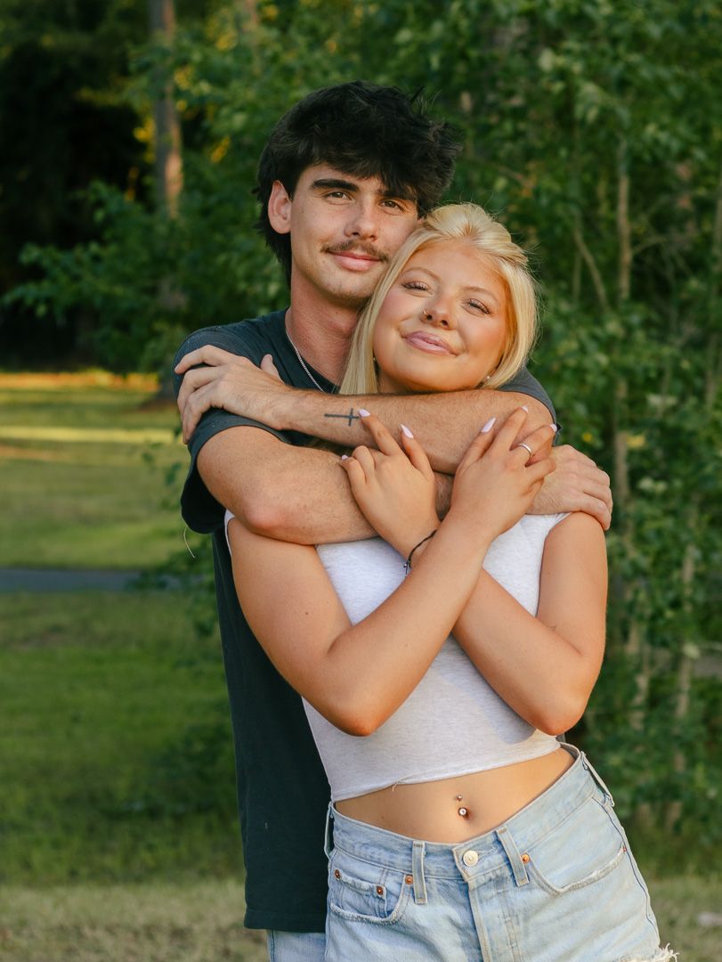 Smiling young couple embracing outdoors, a dark-haired man hugging a blonde woman in a white crop top and jeans with green trees behind.