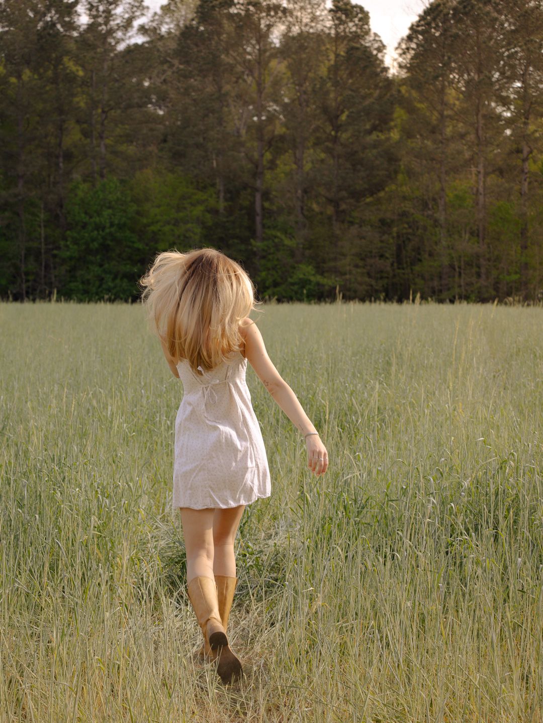 Woman in a white sundress and boots running through a tall green grass field toward a tree line at golden hour.