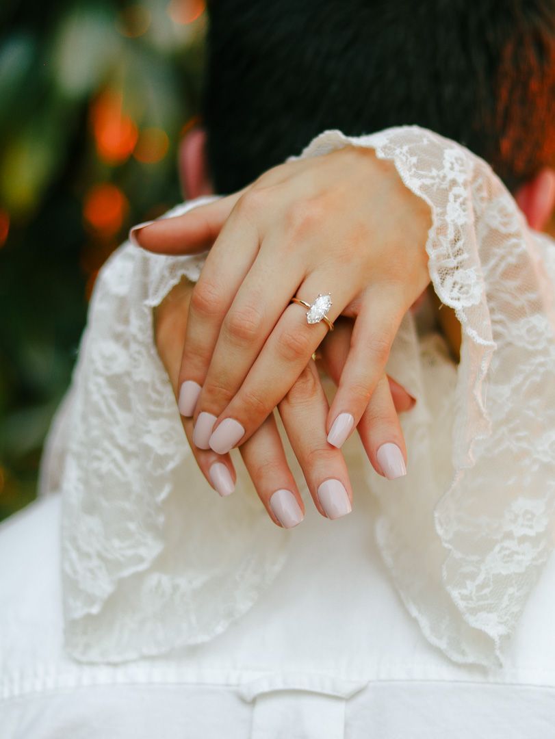 Woman's hands with pale pink manicure crossed over her shoulder, showing an oval diamond engagement ring and white lace sleeves.