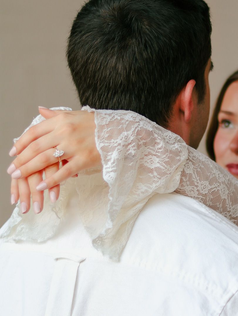 Close-up of a woman's hand with a sparkling diamond engagement ring resting on her fiance's shoulder as they embrace in white clothing.