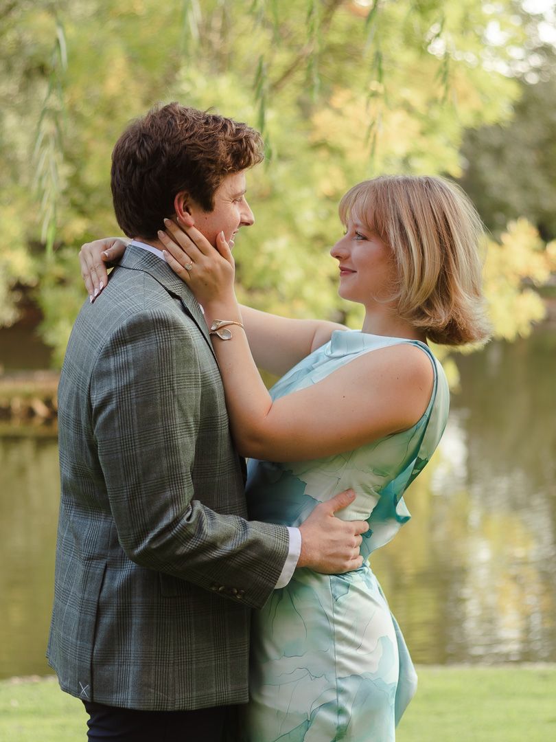 A smiling couple embraces by a tranquil pond beneath weeping willow trees, the woman in a mint dress touching her partner's cheek.