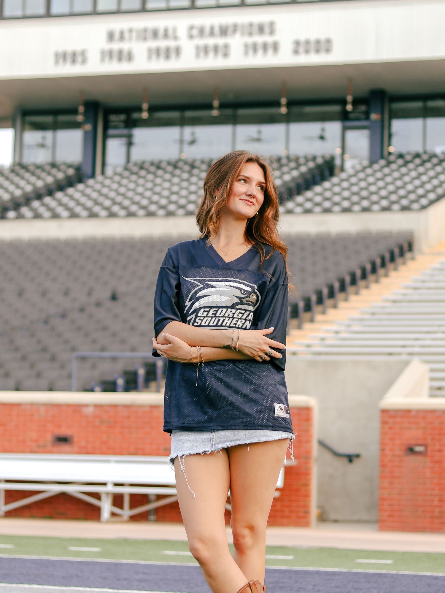 Woman in a Georgia Southern jersey and denim shorts poses with arms crossed in a stadium beneath a national champions banner.