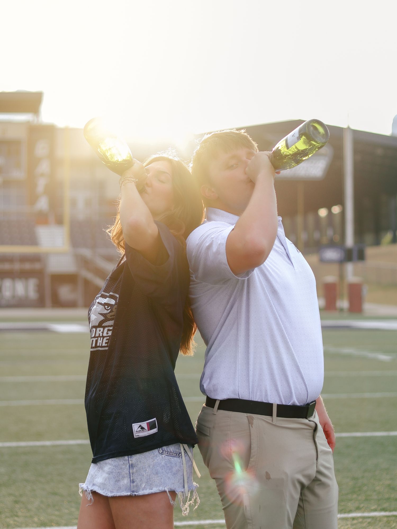 Two people stand back to back on a football field at sunset, each tilting a champagne bottle to their mouth while celebrating.